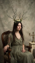 Load image into Gallery viewer, Front three-quarter view of the branch horn headdress with green and cream roses worn by a seated model in a sage green dress in a studio setting (AI-generated model image)