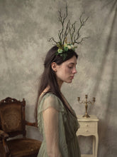 Load image into Gallery viewer, Side profile of the branch horn headdress with green and cream roses worn by a model in a sage green dress in a studio setting (AI-generated model image)
