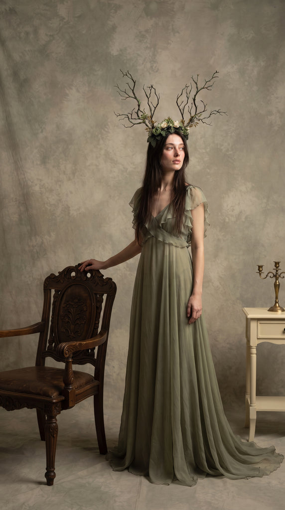Full-body view of the branch horn headdress worn by a model in a long sage green dress standing beside a carved wooden chair in a studio setting (AI-generated model image)