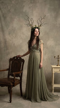 Load image into Gallery viewer, Full-body view of the branch horn headdress worn by a model in a long sage green dress standing beside a carved wooden chair in a studio setting (AI-generated model image)