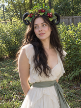 Load image into Gallery viewer, Front three-quarter view of the black ram horns with green leaves and deep red flowers worn by a model with long dark hair in a cream dress outdoors among green bushes (AI-generated model image)
