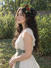 Load image into Gallery viewer, Side profile view of the black ram horn with green leaves and deep red and pink flowers worn by a model with long dark hair in a cream dress outdoors (AI-generated model image)
