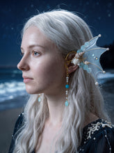 Load image into Gallery viewer, Side three-quarter view of a light blue mermaid fin ear cuff with shells and a dangling pendant worn by a model with long white hair (AI-generated model image)