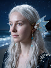 Load image into Gallery viewer, Three-quarter profile of a light blue mermaid fin ear cuff with shells and a pearl and blue bead pendant worn by a model with long white hair (AI-generated model image)